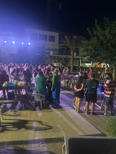 This is a nighttime, medium-wide shot of an outdoor gathering in Blanca, Spain. A crowd of approximately 40-50 people, appearing to be young adults and adults, are socializing and standing around. The scene is illuminated by stage lights from the left, casting a purple and blue glow. In the background, there is a multi-story building with windows and a balcony, and to the right, a palm tree and dense foliage. Several tables are set up with food and drinks, and some people are holding beverages. One individual is dressed in a green character costume, resembling Luigi from the Mario video game series, with blue overalls. Another person in the foreground is wearing a green soccer jersey with "ALONSO 14" on the back. The ground is paved with cobblestones and concrete. The overall atmosphere suggests a festive event or party.