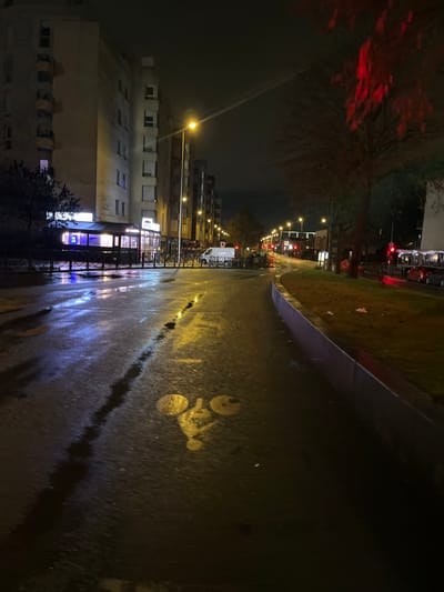 A night street scene with buildings on the left, illuminated by streetlights. The wet asphalt reflects the lights. A dedicated bike lane is marked with a bicycle symbol on the right. Traffic lights and vehicles are visible in the distance, adding to the urban setting. A tree with red leaves is on the upper left.