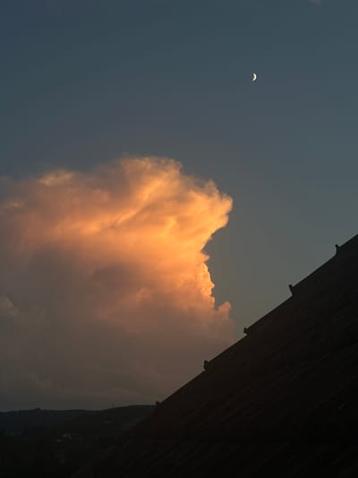 The image captures a twilight scene over Heinebach, Germany, with a prominent, brightly lit cumulus cloud dominating the upper left quadrant. The cloud is illuminated by warm, orange and yellow hues, suggesting it is catching the last rays of the setting sun. To the right of the cloud, a sliver of the crescent moon is visible in the deep blue sky. In the foreground, the dark silhouette of a tiled roof angles across the lower right portion of the frame. The background reveals a dark, silhouetted landscape of rolling hills and trees, with faint lights suggesting a settlement.