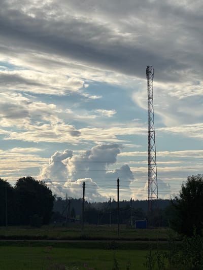 The image depicts a rural landscape under a dramatic sky. A tall, metallic telecommunications tower stands prominently in the mid-right portion of the frame, its structure silhouetted against the clouds. The sky is filled with a mix of dark, heavy clouds and patches of brighter, illuminated sky, creating a sense of dynamic weather. In the foreground, a dark, dense line of trees and vegetation runs across the bottom half of the image, with some power lines extending horizontally through the scene. Further back, more trees form a distant treeline, and several utility poles are visible. The ground appears to be grassy fields. The scene is likely located in the Нёманскі сельскі Савет of Belarus, a rural administrative division.