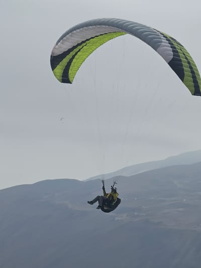 The image captures a paragliding scene, likely over the city of Alto Hospicio, Chile, suggested by the arid landscape in the background. The paraglider is suspended in mid-air, with a green, white, and black striped paragliding wing above. The pilot is visible below the wing, wearing a dark jumpsuit, a backpack, and a helmet. The pilot seems to be maneuvering the paraglider, with hands raised towards the controls. The sky is overcast, suggesting a cloudy day. Another distant paraglider is faintly visible in the background. The landscape reveals dry, mountainous terrain with hazy conditions.