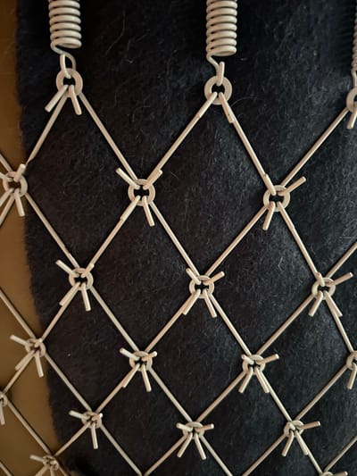 The image shows a close-up view of a diamond-shaped wire mesh. The mesh is made of light-colored metal wire, and each intersection of the wires is secured with a small ring. Some of the wires extend beyond the rings and are bent outwards, forming short spikes. At the top of the image, several coiled springs are attached to the mesh via rings. The mesh is positioned in front of a dark, textured material that appears to be some kind of fabric or felt. The background is a blurry, warm brown color. The image does not depict any specific location in Mlyniv, Ukraine, nor does it contain any people, actions, or interactions.