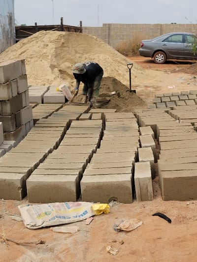 This image depicts an outdoor, unpaved block-making site in the city of Nwofia, Nigeria, under an overcast sky. In the middle ground, a lone individual, appearing male, is bent over. This individual wears a light-colored bucket hat, a dark short-sleeved shirt, and dark trousers, and is actively using a small hand tool, possibly a trowel, to manipulate sand on the reddish-brown earth. Behind the individual, a large mound of light-colored sand is prominent, with a long-handled shovel stuck upright in a smaller pile of sand to the right.
Extending from the foreground into the middle ground are multiple parallel rows of freshly formed, light-grey concrete blocks resting directly on the ground. A partial stack of similar blocks is visible on the far left of the foreground. In the immediate foreground, a partially full or empty BUA CEMENT bag lies on its side, accompanied by a crumpled yellow plastic bag and other minor debris.
In the background, a grey sedan car is parked on unpaved ground to the right of the large sand mound. Behind the car and extending across the scene, a low wall constructed from concrete blocks is visible, partially obscured by sparse dry vegetation. A metal structure is partially visible on the far left of the background.