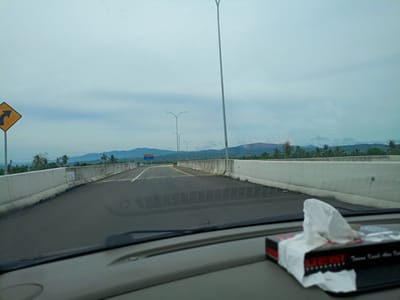 The image shows a view from inside a car driving on a bridge or elevated road in Mukee Gogo, Indonesia. The road is paved and has concrete barriers on either side. There is a sign with a yellow background and a curved arrow pointing left on the left side of the road. A row of light poles lines the road ahead. In the distance, there are mountains under a cloudy sky. The car's dashboard is visible in the foreground, with a box of tissues on it. The windshield and wipers are also partially visible.