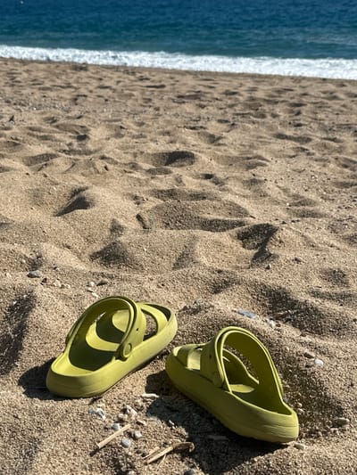 Two lime-green platform sandals are positioned on a sandy beach under direct sunlight in Pineda de Mar, Spain. The sandal on the left faces right, while the sandal on the right faces left, both oriented away from the viewer. The sand, light brown in color, exhibits distinct textures and shadows, with scattered small pebbles and organic debris visible in the immediate foreground. The midground shows more textured sand. In the background, the deep blue water of the Mediterranean Sea is visible, with a white, frothy wave breaking at the shoreline. No individuals are present in the frame.