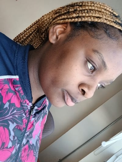 The image is a close-up, profile portrait of a person, initially presented rotated 90 degrees clockwise. The primary subject is an individual with dark skin, brown eyes, and light brown/blonde box braids or similar styled braided hair, which is pulled back.
The person is wearing a zipped jacket featuring a distinctive pattern of pink/magenta, dark blue/black, and gray floral or leaf motifs. A dark-colored garment is visible beneath the jacket. Their face is shown from the side, looking upwards and slightly to their right (when oriented correctly). The expression appears neutral or contemplative.
The background suggests an indoor setting with light-colored walls or a ceiling. A thin, horizontal metal rod or pipe is visible in the upper left section of the frame, and a partial white object with a dotted outline, possibly a hanger or fixture, is also seen on the far left. The lighting is soft and even, indicating an indoor environment without harsh shadows. There is no discernible activity beyond posing for the photograph. No visible text is present in the image. While the location context of Moncton, Canada, is provided, there are no specific visual cues within the image itself to confirm this.