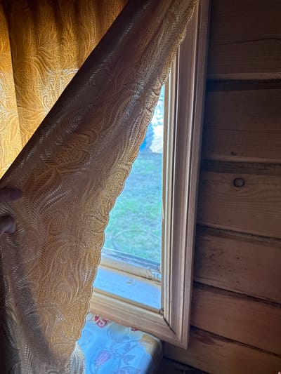 The image depicts the interior of a wooden room, likely a dwelling. A golden-colored curtain with a raised, patterned texture is pulled back to the left, revealing a window. Through the window, a blurred view of a grassy, outdoor area is visible. The window frame is wooden and painted a light color. To the right of the window, the wall is constructed of horizontal wooden planks. A person's finger is visible near the left edge of the curtain. The overall impression is one of a simple, rustic interior.