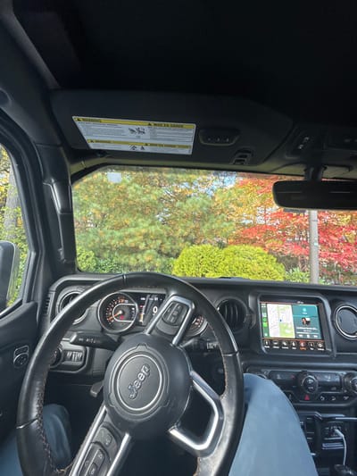 The image depicts the interior of a stationary black Jeep Wrangler, viewed from the driver's perspective. A person occupies the driver's seat, evidenced by a visible leg clad in light blue trousers below the steering wheel. The vehicle's instrument cluster shows a speed of 0 MPH and engine RPMs at 0. The infotainment system displays a map centered on Wellesley, Massachusetts, with "Washington St" visible, alongside active Apple CarPlay media controls. The time is 4:26 PM, and the exterior temperature is 57°F. The front sun visor is lowered, revealing warning labels in English and French. Through the windshield, a vivid autumn landscape is visible, characterized by dense foliage of trees and shrubs exhibiting red, orange, and various shades of green. A utility pole or lamppost is present on the right side of the exterior view. The environment suggests a residential or landscaped area within Wellesley.