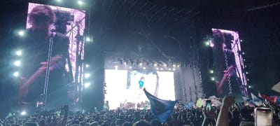 The image depicts an energetic nighttime concert in Tolosa, Argentina, from the perspective of the audience. A large stage structure dominates the background, featuring multiple bright screens and an intricate metal truss system above.
Two prominent vertical screens flank the main stage, each displaying a close-up of a male performer. He has shoulder-length, messy hair and is seen playing an electric guitar and singing into a microphone. The central, wider screen showcases an animated figure wearing a light blue jersey with the number "10" clearly visible on its back, set against a dynamic, abstract blue and white background that resembles water or clouds. Above the stage, a massive, dark circular truss structure is visible against the night sky, adding to the grandeur of the setup.
In the foreground and midground, a dense crowd of people fills the frame, extending far back into the venue. Many audience members are actively engaged, holding up their cell phones to capture the performance. Others are raising their hands or waving flags, including a notable large blue flag in the lower-right foreground. The scene is illuminated solely by the stage lights, creating a vibrant and engaging atmosphere typical of a large outdoor musical event. The time of day is clearly night, as indicated by the dark surroundings. The blue and white flags, combined with the number "10" often associated with Argentine football legends, subtly reinforce the location context.