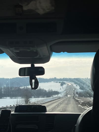 The image, captured from the interior of a vehicle looking forward through the windshield, depicts a winter landscape near Samolet, Russia. The foreground includes the vehicle's dark gray dashboard and an overhead console with various vents and compartments. A rear-view mirror is centrally positioned, from which a dark, rectangular air freshener hangs. No individuals are visible.

The scene outside features a two-lane asphalt road, partially cleared of snow, extending into the distant rolling hills. Snow and slush are present on the road's shoulders and median, and in some areas, partially cover the driving surface. The surrounding landscape consists of snow-covered fields and hills, dotted with bare trees and dense clusters of dark, leafless vegetation. In the middle ground, on the right side of the road, a snow-covered slope parallels the road. Distant structures, possibly small buildings, are faintly discernible on the snowy horizon. The sky is overcast with a bright, hazy appearance, suggesting a cloudy or foggy day.