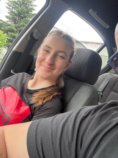 In the city of Vlahovo, Bulgaria, a young woman with light brown hair pulled back in a ponytail, wearing a black t-shirt with a large red graphic, is seated in the back seat of a car. She is smiling and looking at the camera, with her hair falling over her shoulder. A seatbelt is visible behind her. In the foreground, part of another person's arm and shoulder, wearing a black t-shirt, is visible. The car interior, including the headrest of the seat in front of the woman, is also visible. The windows show trees and a building in the background.
