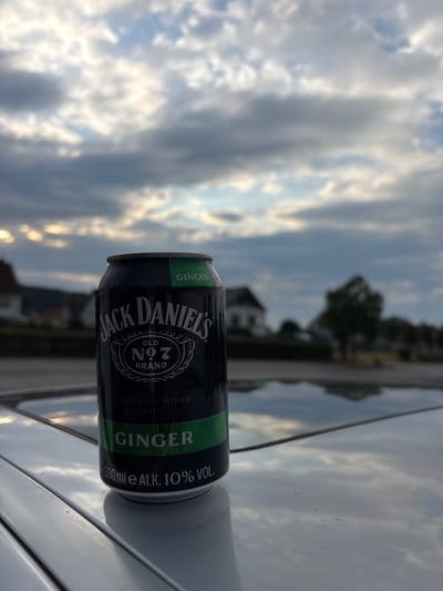 This media file shows a close-up, vertical shot of a black Jack Daniel's can with a green band, resting on a light-colored surface, possibly a car. The can is labeled "JACK DANIEL'S OLD NO. 7 BRAND GINGER" and indicates "330ml e ALK. 10% VOL." The background is blurred but depicts a sky with dramatic clouds, suggesting either dawn or dusk, with some buildings and trees faintly visible, hinting at a suburban or residential setting. The lighting and cloud formation suggest it is late afternoon or early evening. The scene appears to be outdoors, with the can likely placed on a vehicle's surface reflecting the sky. There are no people visible in the image. The overall mood is somewhat atmospheric due to the dramatic sky.