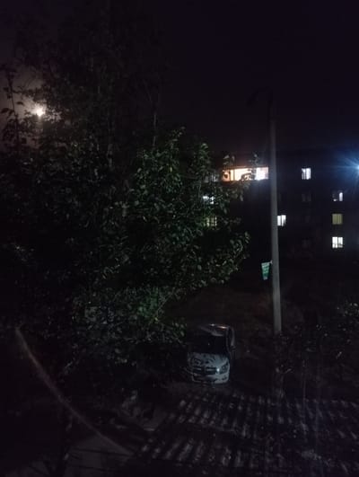 At night in Nurabad, Uzbekistan, a white car is parked on an unpaved area near a large tree and a residential building. The car's headlights are on. The building, a multi-story structure, has several illuminated windows, indicating occupation. A street light is visible, casting a bright illumination to the right of the building. The foliage of the tree is dense and dark, partially obscuring the view of the building and the car. The ground is uneven and appears to be covered with debris. The sky is dark and overcast, with a faint light source in the upper left quadrant, possibly the moon or a distant light.
