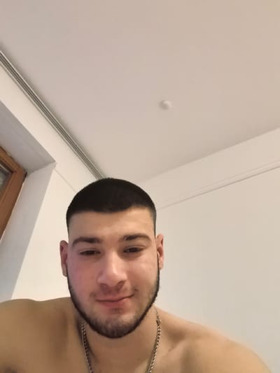 A close-up, slightly low-angle selfie depicts a young man, likely in his late teens or early twenties, looking directly into the camera with a neutral to slightly smiling expression. He has short, dark hair styled in a buzz cut and a dark, trimmed beard and mustache. He is bare-chested, showing his shoulders and upper chest, and wears a silver-colored chain necklace.

The background is dominated by a plain, light-colored (off-white or white) ceiling and wall. On the far left, a portion of a wooden window frame with a glass pane is visible. A metallic or plastic strip, possibly a lighting track or vent, runs along the ceiling near the left wall. A small, circular, white object, which could be a smoke detector or a small light fixture, is present on the ceiling in the upper central area. The indoor lighting appears to be artificial. There is no visible text in the image. While the provided location context is Aystetten, Germany, this detail is not discernible from the visual information alone.
