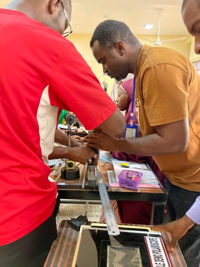 The image shows two men in a room, possibly in Afaka, Nigeria, observing a polariscope demonstration. One man wears a red and white shirt and glasses, and the other wears a tan shirt. A woman in a headscarf is also present. The polariscope is on a table with other items, including a ruler and a purple container. The room appears to be indoors, possibly a classroom or laboratory, with a ceiling fan visible. The men are focused on the demonstration, likely examining the properties of polarized light.