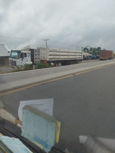 The image depicts a roadside scene in Odiderengwaji, Nigeria, under a cloudy, overcast sky, suggesting it's daytime. The main focus is a white truck carrying several large, cylindrical gas tanks, labeled "power gas". The truck is parked on the side of a road, separated from the camera's vantage point by a concrete barrier.

Next to the truck, a few people are visible, one person wearing a red shirt appears to be attending to something on the truck, while another person is partially visible in the background. Other vehicles, including a blue truck and a brown shipping container on a trailer, are also present further down the road, indicating traffic or a gathering. The background shows buildings with corrugated iron roofs and palm trees, typical of a tropical environment.

The foreground shows a portion of a car's interior, with stacks of papers, possibly documents or a ledger, visible on the dashboard. The overall atmosphere seems calm, with no obvious signs of distress. The weather appears humid and potentially rainy, given the cloud cover. The scene suggests a commercial or logistical operation, possibly involving the transport of gas.