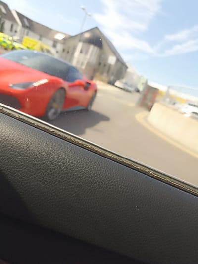 A red sports car is blurred in the background, seen through a car window with a black leather interior. The car appears to be on a road with yellow lines. In the distance, there are buildings and other vehicles, including what looks like an ambulance. The scene is outdoors during the day with a bright sky. The photograph was taken in Dungloe, Ireland.