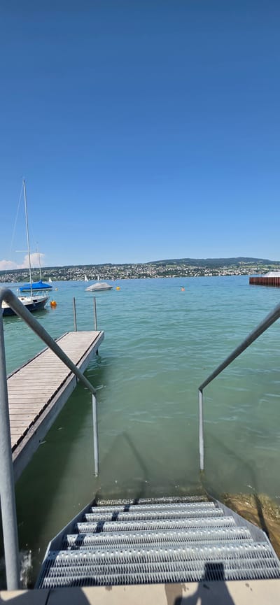 A wide-angle, vertical shot captures a sunny day at the Oberrieden, Switzerland lakeside. In the foreground, a metal staircase with handrails descends into the clear, turquoise water. To the left, a wooden pier extends into the lake, with a sailboat moored to its side. Several other boats are visible on the water, including one covered with a gray tarp and a small speed boat. In the distance, the town of Oberrieden is nestled on a hillside overlooking the lake, with a backdrop of green, rolling hills under a bright blue sky. A few small white clouds dot the horizon. The water is calm, with gentle ripples. The scene suggests a recreational area, likely a public access point to the lake for swimming and boating.