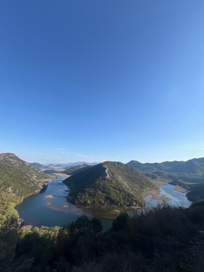 The image captures a scenic view of the Crnojevića River and surrounding landscape in Rvaši, Montenegro. The river winds through a valley surrounded by mountains covered in dense green vegetation. The clear blue sky contrasts with the earthy tones of the terrain. There are no people visible. The perspective is from an elevated viewpoint.