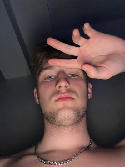 A young Caucasian male with light brown hair and blue eyes is shown in a close-up, low-light selfie. He is wearing a silver chain necklace. His right hand is raised, obscuring the top of his forehead and partially covering his eyes. His left hand is not visible. The background is dark and appears to be an interior space. The image does not provide any information about the location in Vanino, Russia.