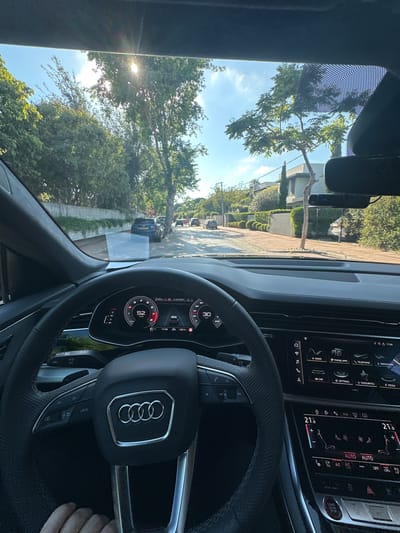 Captured from the driver's seat of an Audi, the image offers a perspective of the residential neighborhood of Kfar Shmaryahu, Israel. The car's dashboard is prominently featured, showcasing the steering wheel with the Audi emblem, digital instrument cluster displaying a speed of 52, and the infotainment system. The climate control settings are visible on the dashboard, set to automatic.

Outside the vehicle, a residential street is lined with trees and houses, typical of Kfar Shmaryahu. A car is visible in the distance. The time displayed on the car's digital instruments is 17:09. The sun is visible through the trees, causing lens flare. A portion of a person's fingers are visible in the bottom right corner of the image.