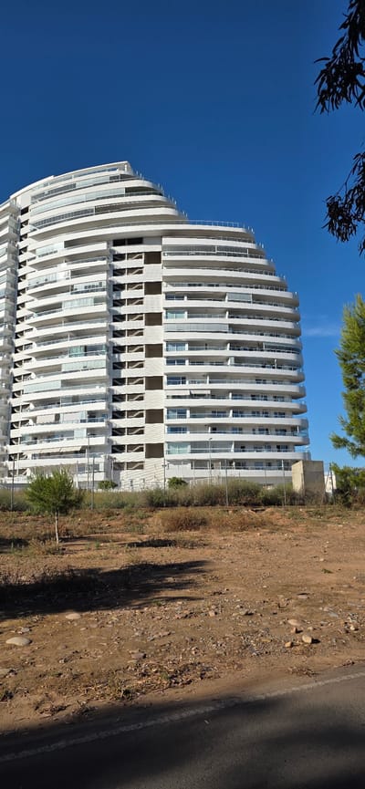 The image shows a tall, white, curved building in Canet d'en Berenguer, Spain. The building has many floors with balconies. To the right of the building is a dirt area, followed by a road with a white line. The sky is blue, and there are tree branches visible at the top of the image.