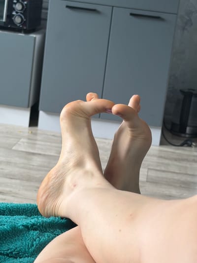 The image shows a close-up view of a person's feet, resting on a turquoise textured surface, possibly a towel or rug. The feet are positioned in a way that their soles are facing upwards and the toes are bent. The background reveals a kitchen or similar domestic environment with modern gray cabinets and a wooden floor. The scene is indoors, and there are no visible indications of the specific location within Старые Туймазы, Russia, beyond it being a residential interior.