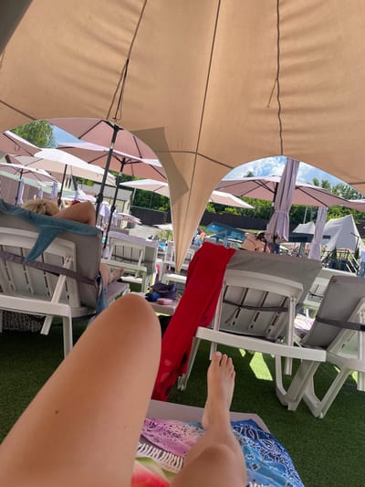 A first-person perspective shot shows a person's legs and feet resting on a lounge chair under a large tan canopy, at an outdoor pool area. The lounge chairs are white plastic with grey cushions, and the ground is covered in artificial grass. In the background, several pink and white umbrellas are set up, and people are seen relaxing and walking around. A blue towel is draped over one of the chairs. The environment suggests a sunny day at a resort or public pool. The location is identified as Zazymia, Ukraine.