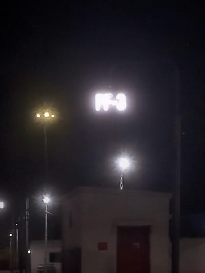 The image shows a nighttime view in what appears to be an industrial or commercial area of Igatpuri, India. A brightly lit sign displaying the characters "H-3" is visible on the left side of the frame. To the right of the sign, several bright streetlights illuminate the scene. A large, light-colored structure with a red section is present on the right side of the image. Additional streetlights are visible in the lower right area, further indicating the location's urban nature. Overall, the scene suggests an area with industrial or commercial activity occurring at night.
