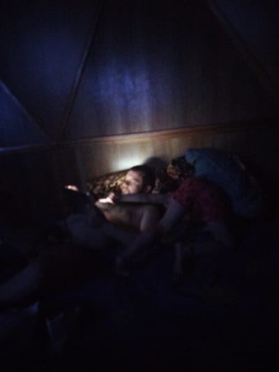An out-of-focus, dimly lit image shows two people lying in bed, possibly in a tent or a room with a triangular ceiling. A faint light source, possibly a phone or tablet, illuminates the face of one person who appears to be looking at it. The other person is partially visible, wearing a red garment. The environment is dark, with only faint outlines of the room and bedding discernible. The specific location in Sovetsk, Russia, cannot be determined from the image alone.