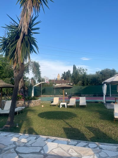 The image depicts a sunny outdoor scene in Ipsos, Greece, with a view of a grassy lawn leading to a tennis/basketball court surrounded by a green fence. White patio chairs are arranged on the lawn. In the background, a building with a golden-colored spire rises above the trees. A palm tree occupies the lower left corner of the image. A stone patio extends along the right edge of the image. Power lines stretch across the clear blue sky above.