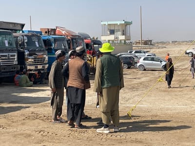 The image depicts an outdoor, dusty vehicle lot in Pir Dust, Afghanistan, under a clear, bright sky. In the foreground and midground, a group of five men is gathered, conversing. One man, seen from behind, wears a neon yellow bucket hat, an olive green vest, and tan traditional clothing. To his left, another man wears a brown vest over black traditional clothing. A third man, to their left, wears a plaid shirt and tan traditional attire, holding a brown object, and appears to be smiling. Additional men are partially visible within this group.
To the left of the main group, a line of large trucks, primarily dump trucks, is parked. Two individuals in orange high-visibility vests are seated on the ground near the front of a dark truck. To the right of the central group, several passenger vehicles, including a silver hatchback and dark SUVs, are parked in an open area. Further in the background, a two-story light blue and white building, resembling a guard post or control tower, is visible, with a tall metal pole adjacent to it.
The ground is unpaved and very dusty, with tire tracks and some white line markings. A yellow rope or strap with metal anchors is stretched across the dusty ground from the foreground towards the midground on the right. In the far background, the terrain appears barren and slightly elevated. Several other individuals are scattered in the background, some walking. Long shadows cast by the people indicate bright sunlight.