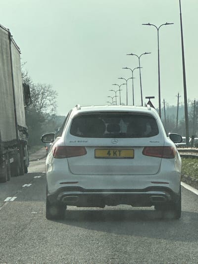 The scene is a major road, likely the A12, within the Brentwood Borough, United Kingdom. A white Mercedes-Benz GLC 300d 4MATIC SUV, bearing the UK registration plate '4 KT,' is viewed from the rear, positioned in what appears to be slowed or stopped traffic. The vehicle features dual exhaust tips and roof rails, with two occupant silhouettes visible through its tinted rear window. To its immediate left, a large grey articulated lorry occupies an adjacent lane, travelling in the same direction. The asphalt road surface exhibits white reflective studs, and a hard shoulder with green vegetation borders the right side. A continuous line of modern streetlights, each with bifurcated heads, extends into the distance along the right. Bare deciduous trees and utility poles are visible in the background under a hazy sky.