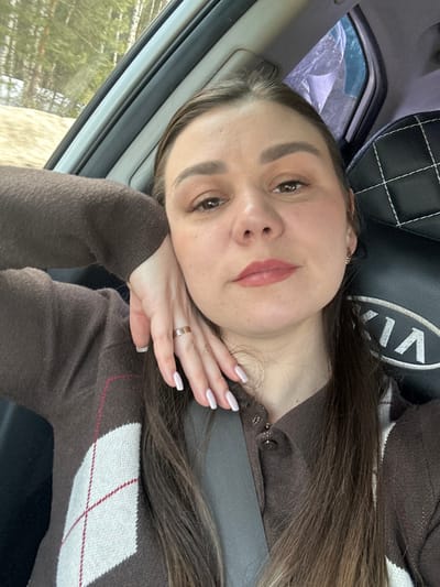 A female individual is depicted in a close-up selfie taken from inside a moving or stationary vehicle, situated in Novoromashkovo, Russia. She has brown hair, dark eyes, and is wearing makeup including light eyeshadow and reddish-pink lipstick. Her left hand, adorned with a gold-colored ring on the ring finger and manicured nails with light polish, rests against her cheek and jawline. She is wearing a brown collared top, possibly a cardigan, featuring a partial white and maroon argyle pattern on the visible left side of her torso. A gray seatbelt extends diagonally across her chest. The vehicle's interior features a black seat headrest with white stitching on the right side, bearing a "KIA" logo. Through the window on the left, an outdoor environment is visible, characterized by tall, thin tree trunks consistent with a forest or wooded area, with patches of snow or light-colored vegetation on the ground, indicating a cool weather season.