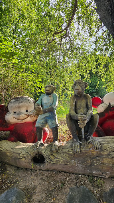 The image shows a collection of weathered concrete statues of monkeys and two large cartoonish figures with red bodies and white faces, arranged on a fallen log. The statues are located outdoors, with lush green trees and foliage in the background, suggesting a park or garden setting. The sun is shining, casting shadows and highlighting the texture of the log and the statues. One monkey statue is standing and wearing blue overalls, while another is sitting and appears to be holding a stick. The cartoon figures have large, round heads and smiling expressions. The ground is covered with dirt, small stones, and fallen leaves. This scene is located at the "Yeonginwon" sculpture park in Cheonan-si, South Korea, known for its collection of whimsical and artistic garden sculptures.