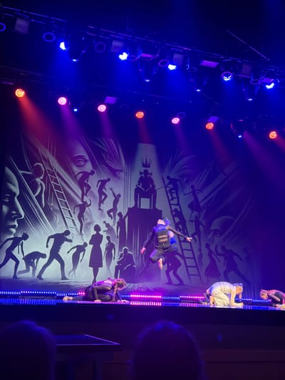 The image displays an indoor stage performance, presumably in Ventspils, Latvia. Six dancers are present on an elevated stage platform. One dancer, centrally positioned, is captured mid-air in a dynamic leap, extending their left arm forward and right leg backward, dressed in a dark, form-fitting costume. Three other dancers are in kneeling or prone positions on the stage floor; two on the left and right wear dark outfits with braided hair, while a central dancer is clad in a light-colored, flowing costume with light hair in a ponytail. Two additional dancers are partially visible on the far right. The stage's front edge is illuminated by parallel rows of blue and pink/purple LED strip lights. Overhead, a complex rigging system supports multiple theatrical lights emitting beams of blue, red, and purple light onto the stage. The backdrop is a large, illuminated screen displaying a monochrome, illustrative graphic. This artwork features numerous stylized human silhouettes engaged in various actions, including climbing ladders, running, and a prominent figure seated on a throne under a crown symbol. Large, abstract faces are also integrated into the background imagery, contributing to a surreal thematic setting. The heads of two out-of-focus audience members are visible in the extreme foreground, indicating the perspective of the shot.