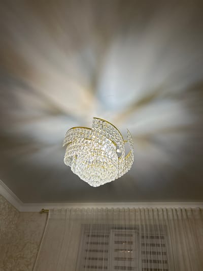 A chandelier hangs from the ceiling, casting a patterned shadow onto the white ceiling. The chandelier is made of clear crystals and gold-colored metal. Below the chandelier, sheer white curtains are drawn across a window with blinds. The walls are decorated with a textured wallpaper. The scene appears to be indoors, possibly in a residential setting in Nizhniye Kazanishchi, Russia. There are no people or animals visible in the image.