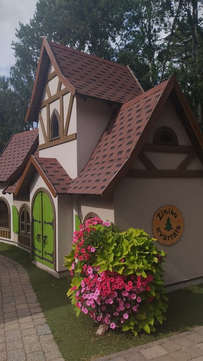 The image shows a charming, fairytale-style miniature house situated in Kroņauce, Latvia. The house features a reddish-brown shingle roof with prominent dark brown trim and decorative wooden beams against a cream-colored facade. Two arched green doors with decorative black hardware are visible on the left side of the structure. Adjacent to the house, a vibrant bush overflows with pink and purple flowers, intermingled with bright green foliage. A circular wooden sign with the inscription "Zinību kvartāls" (which translates to "Knowledge Quarter") and a stylized green tree is mounted on the wall to the right of the flowers. A cobblestone path leads away from the house and flowers towards the left side of the frame, bordered by lush green grass. The background is dominated by the dense foliage of tall trees, suggesting a natural, park-like setting. The sky above is partly cloudy. There are no people or animals visible in the image.