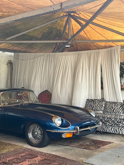 The image shows a blue vintage car parked indoors under a tent-like structure, possibly a garage or event space, in Kraaifontein, South Africa. The tent has a metal frame with a tan fabric roof. There is white fabric draped as a backdrop behind the car, and zebra-patterned cushions and a red chair are visible. A patterned rug sits adjacent to the car. The scene suggests a possible display or event setting for the vintage car.