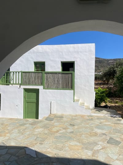 The image captures a static, sunlit exterior scene in Πλατύς Γιαλός, Greece, framed by a grey, arched architectural structure in the immediate foreground. The primary subject is a two-story, whitewashed building featuring green-painted wooden architectural elements.
On the ground level of the building, a central, vertically paneled green wooden door is present. To the right of this door, a five-step, whitewashed staircase ascends along the building's facade. The upper level includes a balcony on the left with a green wooden railing, which incorporates a section of vertical, light-colored bamboo or reed screening. A dark-colored chair is partially visible on this balcony. Two rectangular openings are on the upper facade: one on the left with a green frame and dark panes, and another larger, dark opening on the right.
The ground directly in front of the building is paved with irregular, flat flagstones in shades of grey, beige, and tan, with a shadow from the foreground arch obscuring the bottom portion. The background features a sloping, vegetated hillside to the right of the building, sparsely covered with green shrubs and olive trees. A distant, higher landmass is visible on the horizon. The sky overhead is clear and a vibrant blue. No individuals are present in the scene.