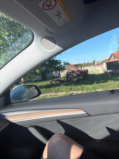 This media file captures an outdoor scene through the windshield and side window of a vehicle, likely a car. The primary visible objects outside the car are a red tractor and a building with an orange roof, suggesting a rural setting in Radko Dimitrievo, Bulgaria. The tractor appears to be an older model, and it is situated on a grassy area next to the building. Trees with green foliage are also prominent, some close to the car and others further in the background. The sky is a clear, bright blue, indicating a sunny day.
Inside the vehicle, parts of the interior are visible, including the car's sun visor with an airbag warning sticker, a speaker, a rearview mirror, and a wooden trim detail. A person's leg, illuminated by sunlight, is visible on the right side of the frame, suggesting the photo was taken from the passenger seat or back seat of the car. The overall impression is a snapshot of a casual moment, possibly during a road trip or a stop in the countryside. The time of day appears to be midday due to the bright sunlight and clear sky.