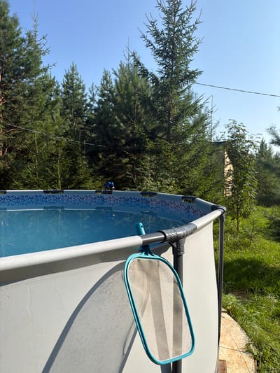 In Kuliki, Russia, a round above-ground swimming pool filled with clear blue water stands in a grassy area surrounded by tall pine trees. The pool has a white exterior and a decorative blue tiled liner visible at the top. A blue and gray pool skimmer is attached to the side of the pool, resting on the edge. The sky is clear and blue.