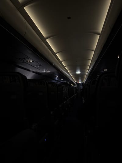 The image depicts the interior of what appears to be an aircraft cabin. On the left, the ceiling and upper sidewall are visible, illuminated by integrated light panels. The opposite side of the cabin shows rows of passenger seats in darkness. An emergency exit sign can be seen at the far end of the cabin. The specific location in Paiania Municipal Unit, Greece, is not determinable from the image alone.