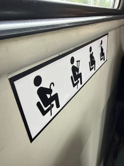 A close-up shot shows a white sign with black icons pasted on a light-colored wall, likely inside a public transportation vehicle. The sign displays four icons: the first shows a person sitting with a cane, indicating priority seating for the elderly or disabled. The subsequent icons depict a pregnant woman, and a person sitting, with the last icon partially obscured. The wall appears to be a textured, slightly worn surface. Above the sign, a dark, horizontal metal bar is visible, running along the top edge of the frame, suggesting the interior of a bus, train, or similar vehicle. The background is out of focus but hints at natural light coming from a window. The image does not provide any direct information about the city of Aprelevka, Russia.