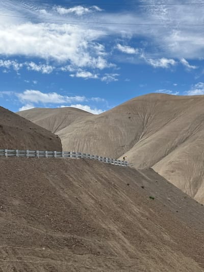 A solo figure on a bicycle is seen on a mountain road in Wakha, India. The road is lined with a metal guardrail on the left side. The surrounding environment consists of arid, barren mountains with visible erosion patterns. The sky above is blue with scattered white clouds, and power lines are visible in the upper portion of the image.
