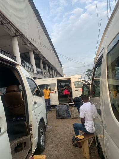The image shows a street scene in Elelenwo, Nigeria, with several white minibuses parked on a dirt and gravel surface. In the foreground, the side of a white minibus is visible with its door open, revealing brown seats and a black fabric covering. To the right, another white minibus has its door open, and a man in a yellow t-shirt and dark pants is standing nearby. Inside this minibus, a woman in a pink dress is seated. Further to the right, a man in a white t-shirt and blue jeans is sitting on a wooden bench next to a silver minibus, eating from a plate. Luggage, including striped bags and a backpack, is scattered on the ground. In the background, a large, multi-story building with columns and balconies is visible. The sky is partly cloudy.