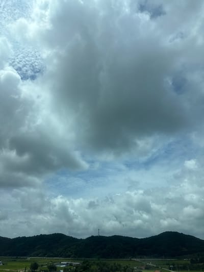 The image depicts a wide shot of a rural landscape in Ungok-ri, South Korea, under a cloudy sky. The foreground features a stretch of green fields, some of which appear to be cultivated with crops. Several low-lying buildings, possibly greenhouses or farm structures, are visible amidst the fields. Roads and utility poles are scattered throughout the area. In the middle ground, a dense, dark green forested hill rises, its contours softened by the distance. A telecommunications tower stands prominently on the crest of the hill. The background is dominated by a vast, overcast sky filled with a mixture of heavy, dark clouds and patches of lighter, more diffused clouds. There are no people or animals visible in the image.