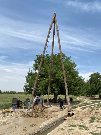 The image depicts a scene in a rural area, potentially near the city of Ashoqah, Afghanistan, given the geographical context. A tall, wooden derrick stands prominently in the foreground, likely used for drilling a well. Several individuals, dressed in traditional Afghan clothing, are gathered around the base of the derrick, appearing to be involved in the well-drilling operation. A large pile of excavated earth sits nearby. A small, engine-powered pump is positioned to the left of the derrick. An irrigation ditch runs along the bottom of the frame, containing murky water and sections of concrete piping. In the background, a field of crops extends toward the horizon, and trees offer shade to additional people who sit under them. The sky is partly cloudy.