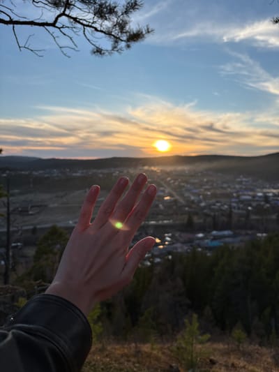 A human hand reaches out toward a low sun partially obscured by clouds in Невон, Russia. The background shows a town and forest.