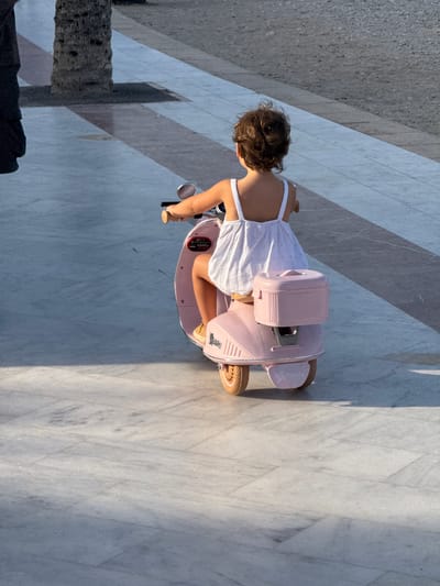 A young girl with curly brown hair is riding a pink toy Vespa scooter on a marble-tiled promenade. She is wearing a white sundress with a floral pattern and tan slip-on shoes. The scooter has a pink storage box on the back and "Twinty" is visible on the rear fender. In the background, a palm tree trunk is visible to the left. The promenade is paved with light-colored marble tiles, with darker grey tiles forming a diagonal pattern in the background. The scene is illuminated by sunlight, casting shadows. The location is Pla Parcial Platja, Spain.